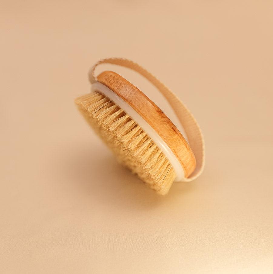 A natural bristle dry brush with a wooden handle, designed for exfoliation and lymphatic detox, placed on a clean, neutral background.